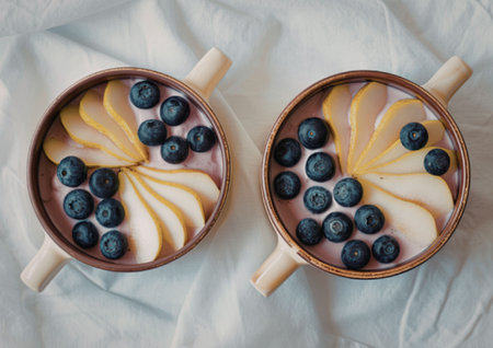 Two brown bowls filled with a pink oatmeal or yogurt, topped with sliced pears and blueberries, placed on a white fabric background.の写真素材