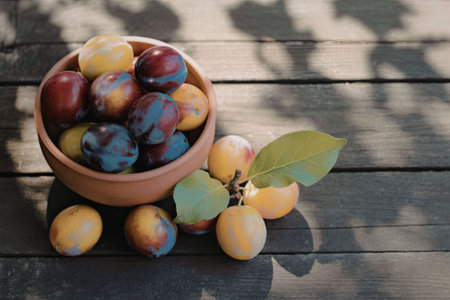 A terracotta bowl filled with a mix of ripe and unripe plums, placed on a dark wooden table with scattered plums and leaves.の写真素材
