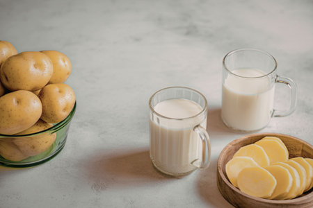 A bowl of yellow potatoes, a bowl of sliced potatoes, and two glasses of potato milk on a white marble surface.の写真素材