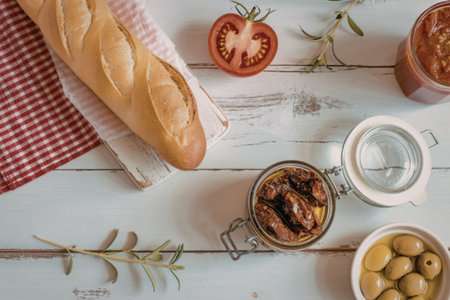 A crusty baguette on a red checkered cloth, accompanied by sun-dried tomatoes, green olives, and fresh rosemary on a distressed white wooden table.の写真素材