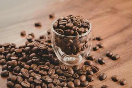 A clear glass cup filled with roasted coffee beans, surrounded by scattered beans on a wooden surface.の写真素材
