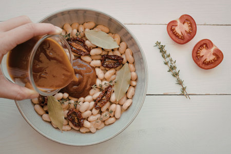 A hand pouring a rich brown sauce into a bowl of beans, sun-dried tomatoes, and herbs, with fresh tomato halves and a sprig of thyme on the side.の写真素材