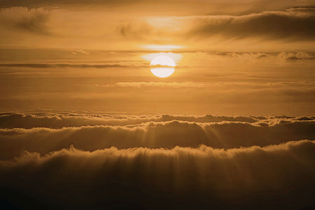A serene sunrise above the clouds with the sun peeking through the gaps, casting a warm glow on the cloudy landscape.の写真素材