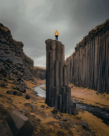 A dramatic landscape featuring a solitary, tall, columnar rock formation surrounded by similar formations and a stream, under a cloudy sky.の写真素材