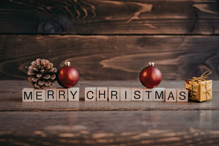 A festive Christmas arrangement on a wooden table with red ornaments, a pinecone, and a gift box spelling out Merry Christmasの写真素材