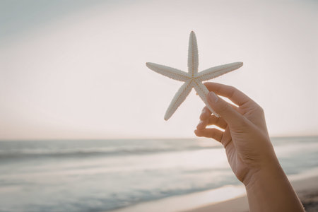 A hand holding a starfish in front of a beach scene with the sun shining brightly.の写真素材