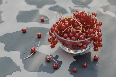 A glass bowl filled with fresh red currants on a gray surface with scattered currants