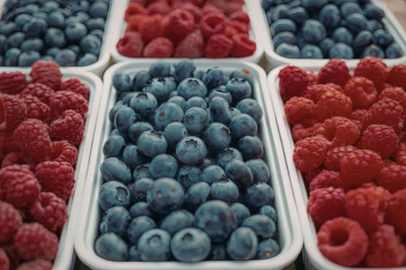 A close-up view of fresh blueberries, raspberries, and more berries in white containers, showcasing...