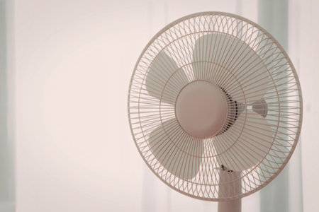The image features a beige standing fan with a mesh grill and three blades, set against a white...