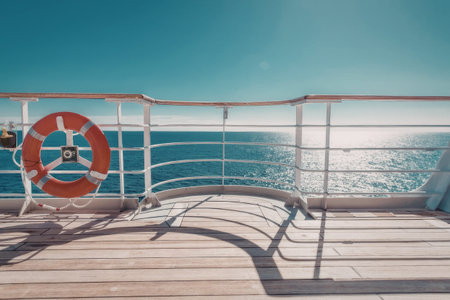 a wooden deck of a boat with a white railing and a red life preserver hanging on it. the ocean is visible in the background with a clear blue sky.