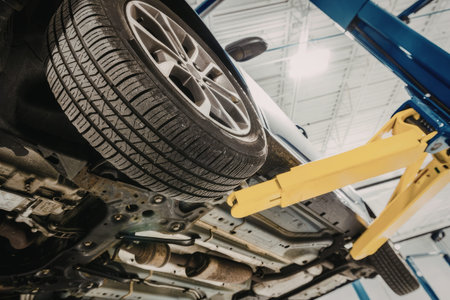 The image shows a car being lifted on a yellow hydraulic lift in a garage. The car is positioned on its side, with the tire and wheel visible. The car's undercarriage is exposed, revealing various components such as the exhaust system and suspension. The background of the image features a white ceiling with metal beams and a blue metal structure that appears to be part of the lift or the garage's framework.