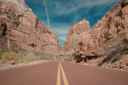 a road with double yellow lines running down the center, surrounded by large red rock formations on either side. the road is paved and appears to be in good condition. the rock formations are tall and have a reddish-brown color, with some greenery growing on them. the sky above is blue with some wispy clouds.