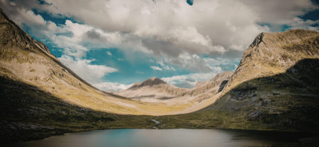 a picturesque landscape featuring a calm lake in the foreground, nestled within a valley surrounded by rugged mountains and hills covered in greenery, set against a backdrop of a blue sky with white clouds