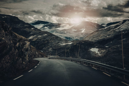 a long, winding road cutting through a rugged mountain landscape. the road has a metal guardrail on the right side and white markings on the asphalt. it curves gently to the left in the distance. the mountains are rocky and steep, with patches of snow visible on the peaks and slopes. the sky above is cloudy and overcast, with the sun peeking through the clouds, casting a warm glow on the scene.