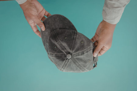 An image of two hands holding a gray baseball cap against a teal background.