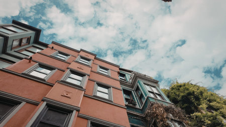 A low-angle shot of a red apartment building with multiple windows and balconies, set against a backdrop of a cloudy blue sky and green trees.