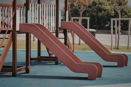 The image shows a close-up view of two red slides on a playground. The slides are made of metal and have a curved shape. They are placed on a blue rubber surface, which is likely designed for safety. In the background, there are trees and grass, suggesting that the playground is located in a park or other outdoor setting.