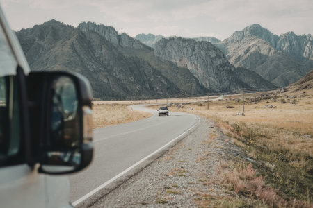 A winding road cuts through a picturesque landscape of mountains and grasslands, with a vehicle's side mirror in the foreground.