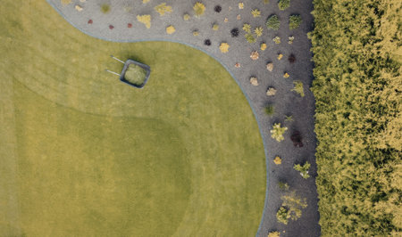 This aerial image shows a vibrant green lawn with a curved edge, adjacent to a garden filled with various plants and a dense line of trees. The lawn is neatly trimmed, and a small, square-shaped object sits on the grass near the garden. The garden itself features a mix of small plants and flowers, adding color and texture to the scene. The trees on the right side of the image are densely packed, creating a natural boundary.