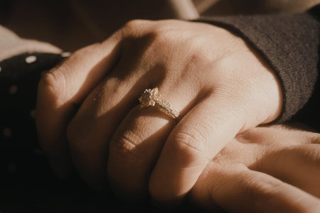 A close-up photo of two hands with one hand gently placed on top of the other, showcasing a diamond engagement ring on the ring finger.