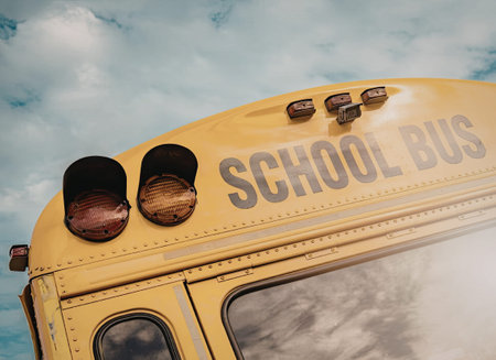 The image shows a close-up view of the top of a yellow school bus with the words 'SCHOOL BUS' written on it in large gray letters. The bus has two red lights on the left side and three smaller lights on the right side. The background is a blue sky with white clouds.