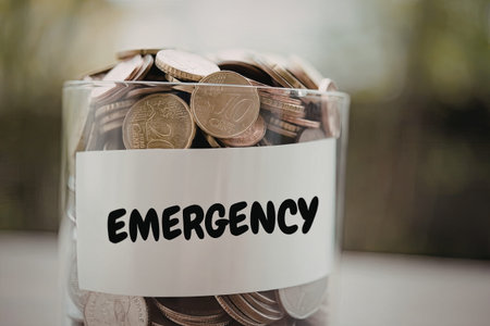 A clear glass jar filled with coins and labeled 'EMERGENCY' for savings