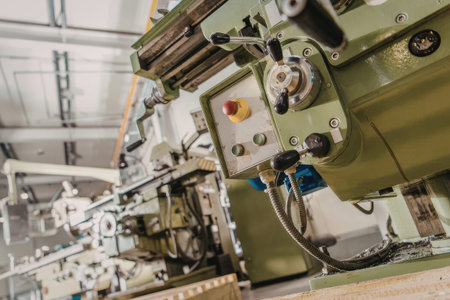 a green industrial machine with a control panel featuring a red and yellow button, a silver dial, and various black handles and wires, situated in a factory setting with other machinery in the background