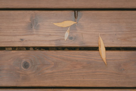 the image shows a close-up of a wooden surface, possibly a deck or a bench, with three yellow leaves scattered on it. the wood is stained brown and has a natural grain pattern. the leaves are dry and appear to be from a deciduous tree. the overall atmosphere suggests a autumnal setting.
