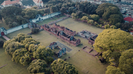 aerial view of the st thomas church ruins in malacca, malaysia. the old church is surrounded by trees and green grass with a white wall and buildings in the background.