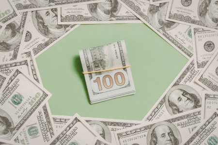 A stack of hundred dollar bills secured with a yellow band on a green octagonal background, surrounded by scattered dollar bills.