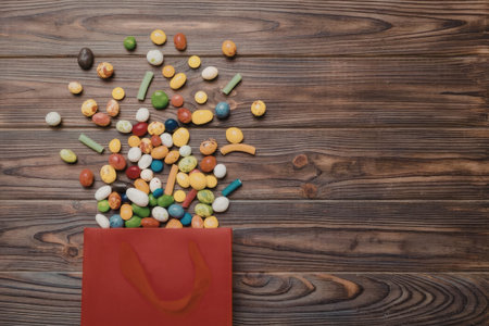 A red paper bag with colorful candies spilling out onto a wooden surface