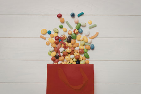 A red bag with a handle, filled with a variety of colorful candies, spilling out against a white wooden background.