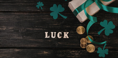 A gift wrapped in paper with a green ribbon and surrounded by shamrocks and gold coins on a wooden background with the word 'LUCK' written in wooden letters.