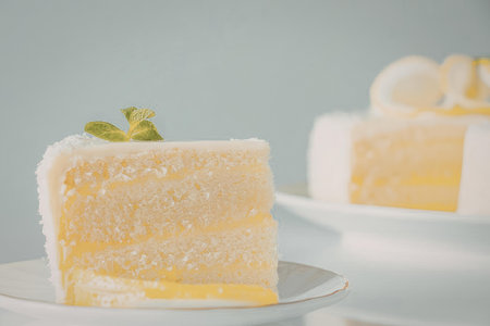 A slice of lemon cake on a plate with a blurred background of another slice of cake.