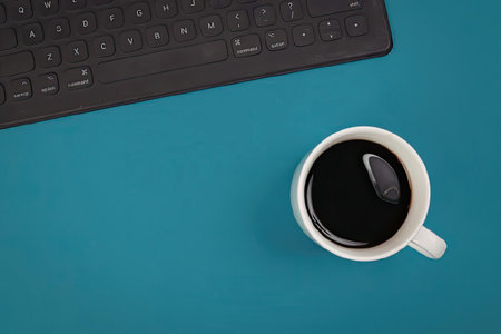 A white coffee cup filled with black coffee sits on a blue desk next to a black keyboard.