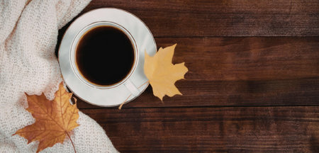 A cup of coffee on a saucer, accompanied by autumn leaves and a blanket on a wooden table.