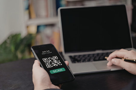 A person is holding a phone with a QR code on the screen and a laptop in the background.