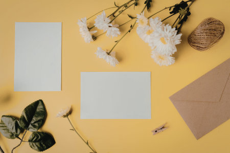 A flat lay of stationery items including two white cards, a brown envelope, white flowers, and green leaves on a bright yellow background.