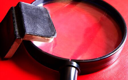 Small black book lying on a big magnifying glasson a red backgroundの写真素材