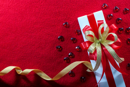 White gift box with red and gold ribbon, and a small red hearts shape on a red velvet floor with copy space, flat lay, Valentine's day background.の写真素材