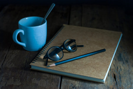 A glasses and a black pencil are placed on a notebook on a rustic wooden floor, with a coffee mug...