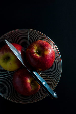 Red apples with a kitchen knife put on a stainless steel round basket on a black background, top view.の写真素材