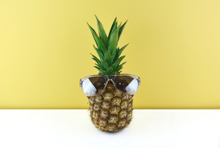 Ripe and juicy pineapple wearing stylish mirrored sunglasses on a white wooden table and pink...