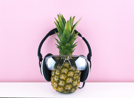 Ripe and juicy pineapple wearing stylish mirrored sunglasses and earphones on a white wooden table and pink background. Place for your text.の写真素材
