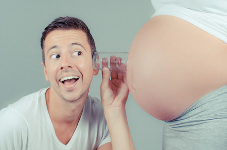 funny father listening with a glass on the belly of his pregnant wifeの写真素材