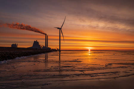A colourful sunrise at Oresund with a powerplant and a modern Windmill close to the shore. The water is covered with ice reflecting the colours.の写真素材
