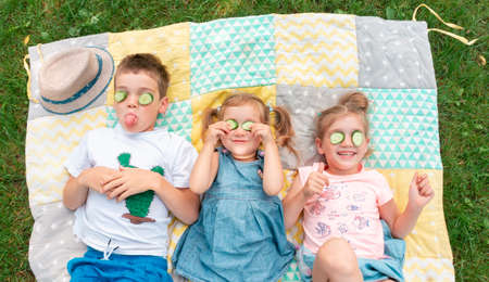 Kids smile and indulge lying on a patchwork rug. Children fooling around with a cucumber mask. The boy shows his tongue. Family summer camp activity ideas. Happy kids spend their summer holidays.の写真素材