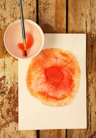 Watercolor blot on paper and a brush in a cup of water on wooden tableの写真素材