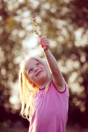 Cute little blonde girl with a flower in her hand on the meadow in spring dayの写真素材
