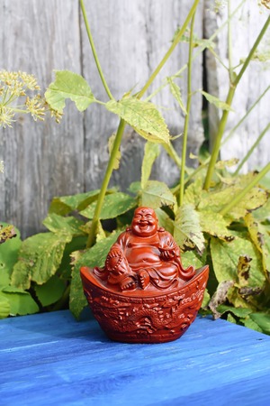 Ceramic buddha figurine on blue aged wooden boards in summer park with flowers.の写真素材